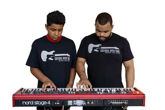 Two males wearing black 'Guitars Over Guns' t-shirts focused on playing a red Nord Stage 4 keyboard together.